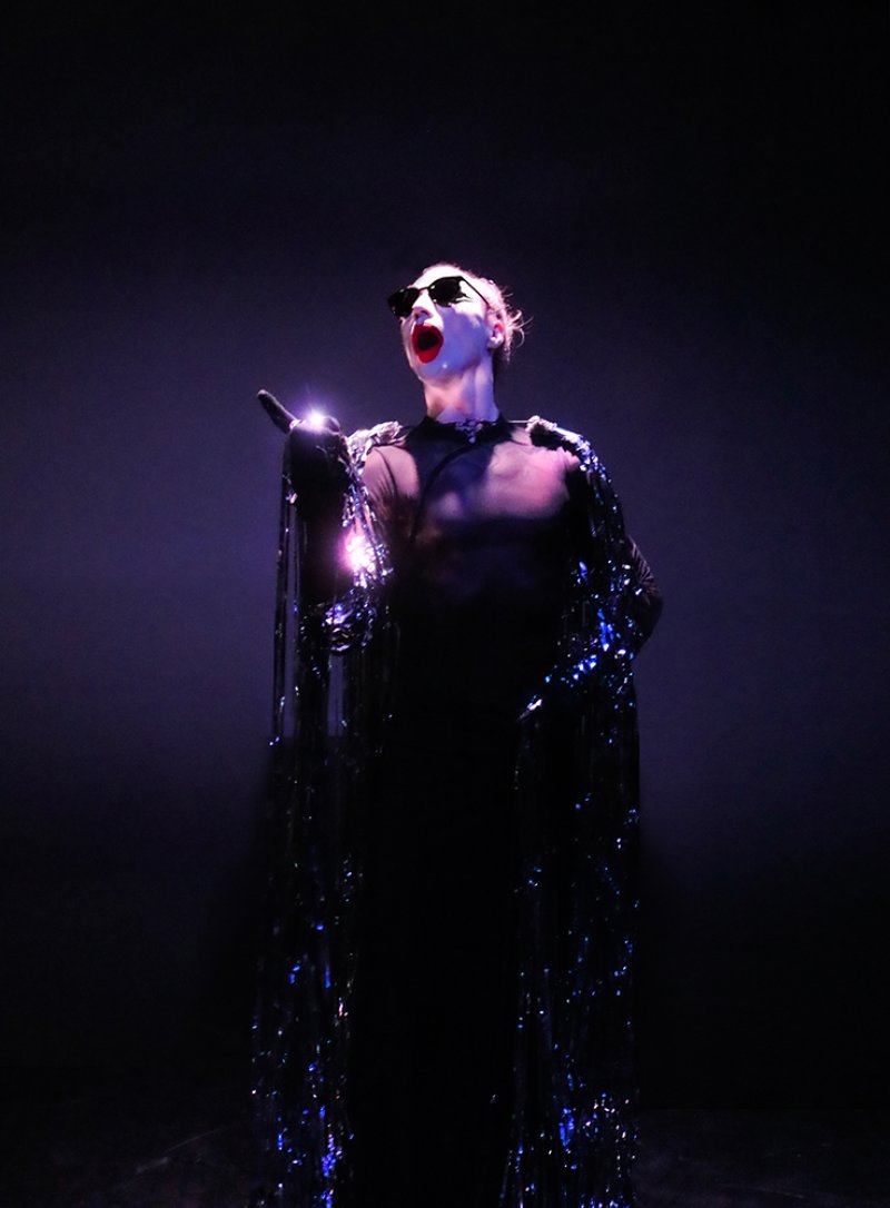 Performer in sunglasses and a shimmering tinsel cape, mouth open with bright red lipstick, against a dark purple background.