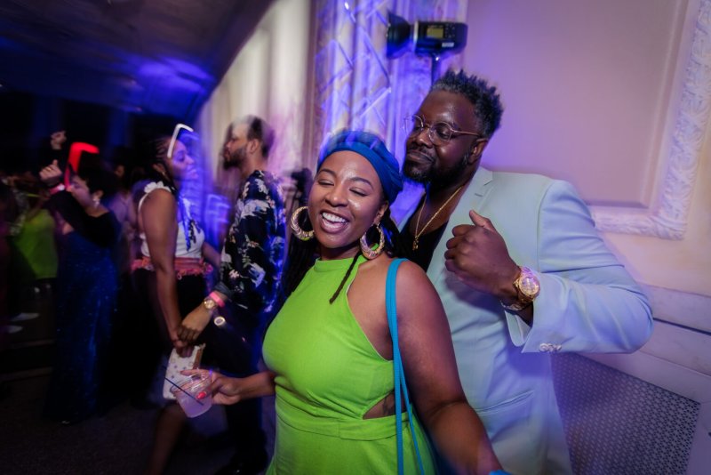 A vibrant house party scene with a Black woman in a lime green dress and blue headwrap laughing, embraced by a Black man in a light blue suit.