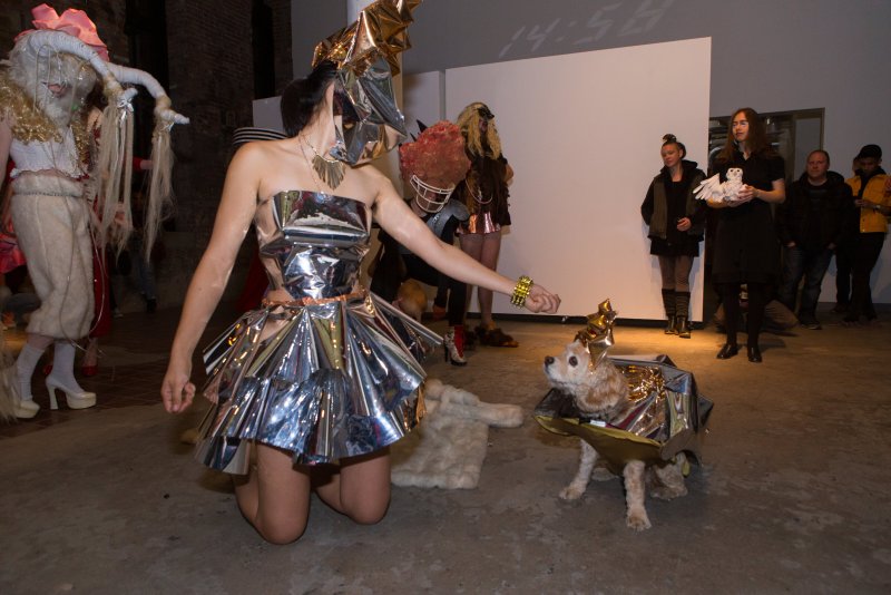 Performance art with a woman in a silver dress kneeling, extending hand to a dog in a matching costume.