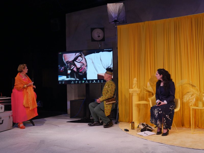 Three people on a stage with a large screen displaying a man's face, against a yellow curtain backdrop.