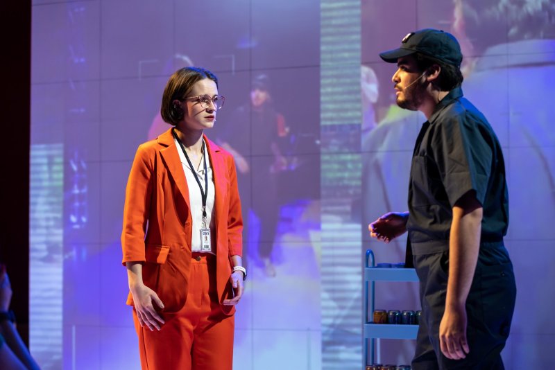 Two figures in a stage setting. A woman in an orange suit faces a man in a dark uniform and baseball cap. A blue-toned projection with grid patterns is visible in the background.