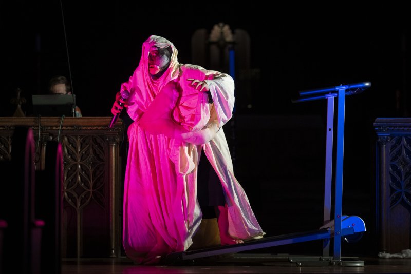 Performer in white and pink robes with black and gold face paint, holding a microphone, standing on a treadmill on a dark stage.