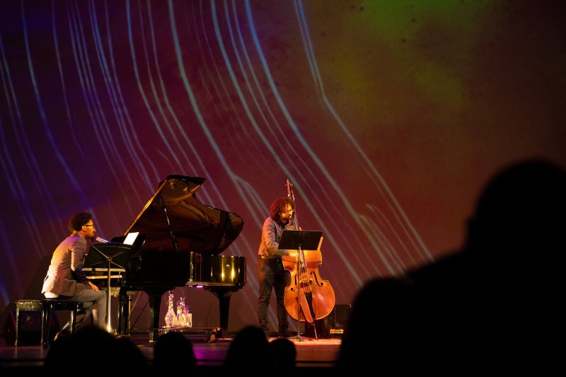 Two musicians, one at a grand piano and another with a double bass, perform on a stage with a purple and red abstract light projection.