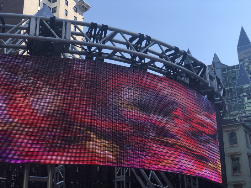 Large outdoor LED screen displaying abstract patterns in red, purple, and yellow. Metal support structure visible above. Buildings and blue sky in background.