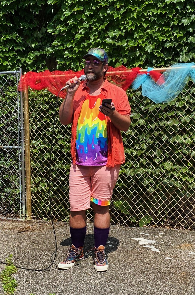 Man in rainbow shirt, pink shorts, and cap speaking into a microphone, holding a phone, in front of a chain-link fence.