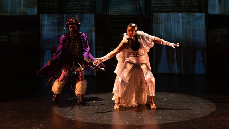 Two figures in costume on a stage, illuminated by spotlights. One wears purple and the other white.