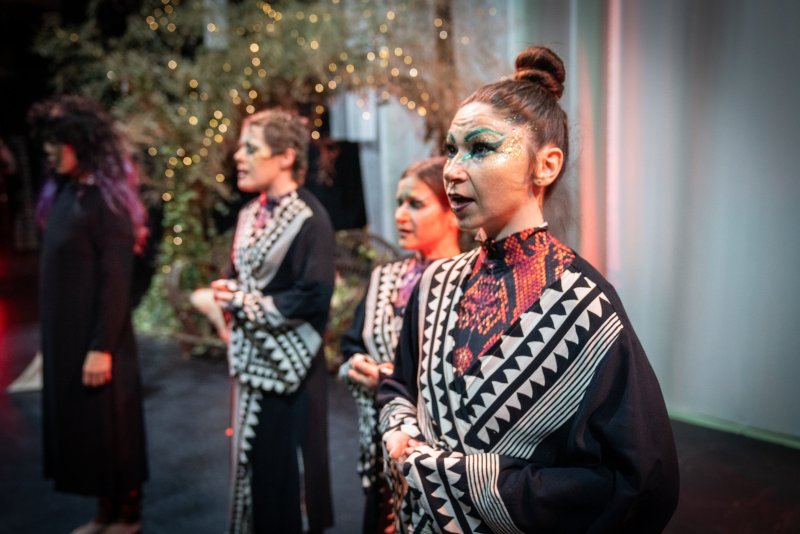 Woman with elaborate face paint and bun, wearing a black robe with geometric patterns, stands among other costumed figures.