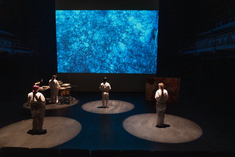 Four performers in white stand in spotlights on a dark stage, facing a large screen displaying blue bubbles.