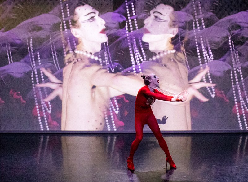 Performer in red bodysuit and heels on a dark stage, with two mirrored figures projected behind them.