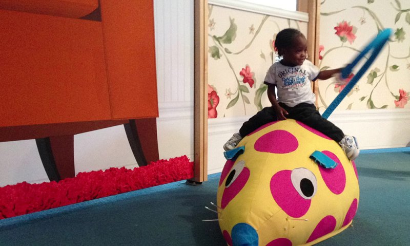 Installation featuring a child riding a yellow, pink-spotted, plush toy. A large orange block with red fringe sits to the left, set against floral wallpaper. Blue carpet grounds the scene.