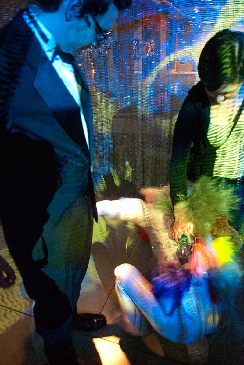 Two people in a dark room with blue and green light, one standing, one kneeling, surrounded by colorful, blurry objects.