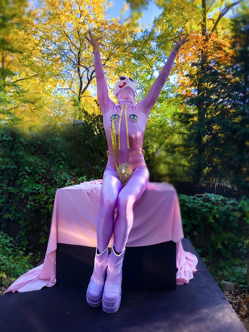 Figure in a lavender bodysuit with eye motifs and platform boots sits on a pink-draped table. Arms are raised against a backdrop of green foliage and yellow-leaved trees.
