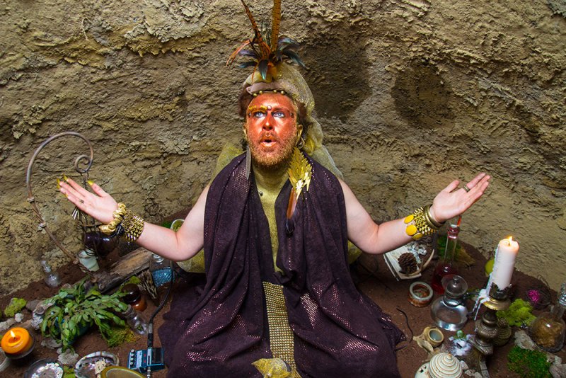 Person with gold painted skin and feather headdress sits in a cave with arms outstretched. Candles, plants, and vials surround them. They wear a dark shawl and gold jewelry.