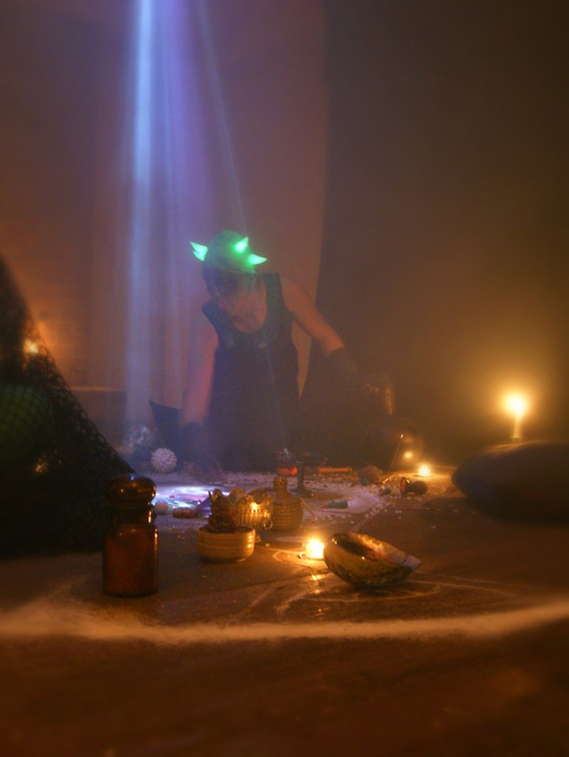 A person with glowing green horns kneels in a dimly lit room, surrounded by candles and ritualistic objects.