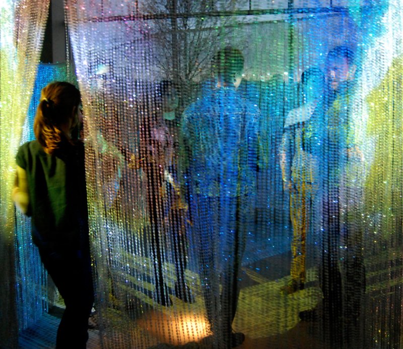 People stand behind a shimmering curtain of colorful beads, illuminated by blue, green, and yellow light.