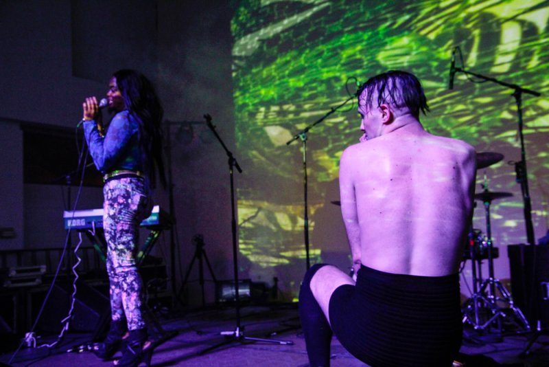 Performance scene with singer, keyboard, drums, and shirtless performer kneeling; green projection on wall. Purple lighting.