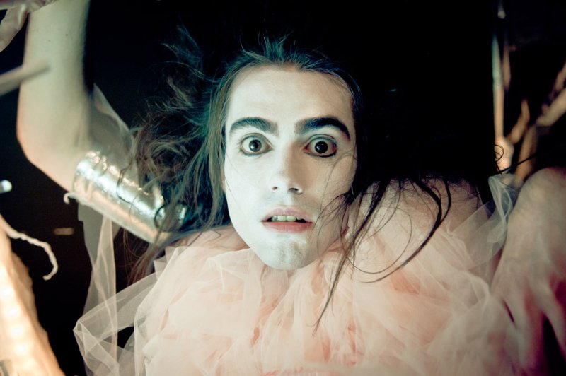 Performer with white face paint, dark messy hair, wide eyes, and a ruffled pink tulle collar, looking up.