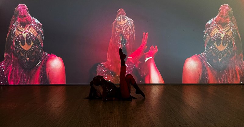 Performer in dark costume on a wooden floor, with three projected images of a person in a red, sequined outfit on a dark wall.