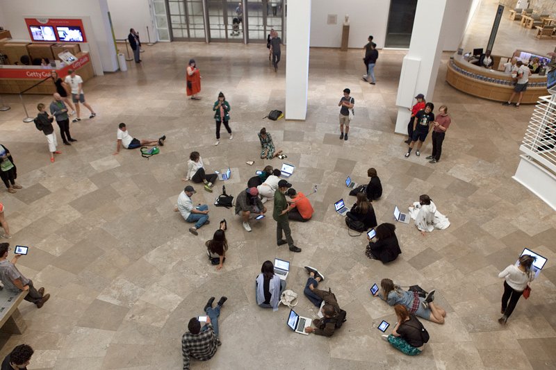 Overhead view of a large, light-tiled gallery space with many people sitting on the floor using laptops and tablets.