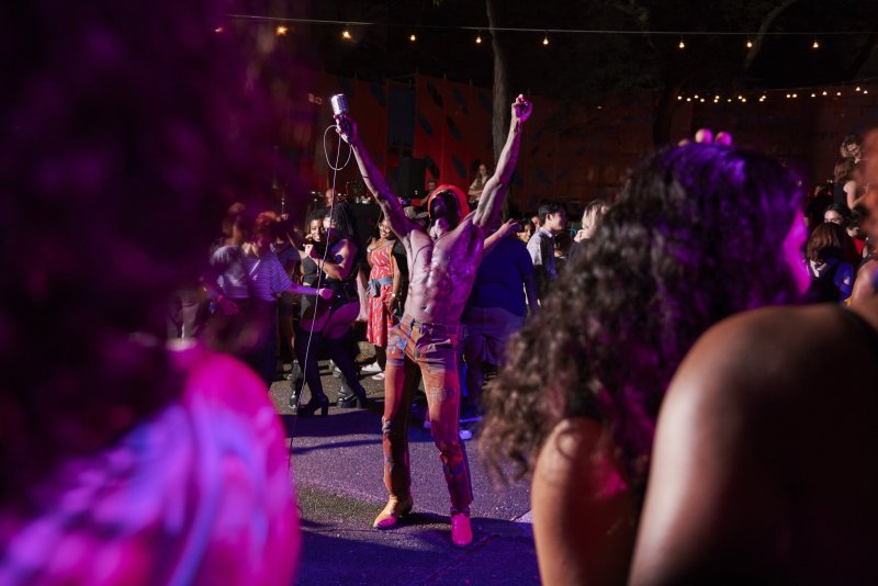 Performance scene with a shirtless person holding a microphone, arms raised, surrounded by a crowd. Purple and pink lighting illuminates the scene. Red and blue backdrop with hanging lights.