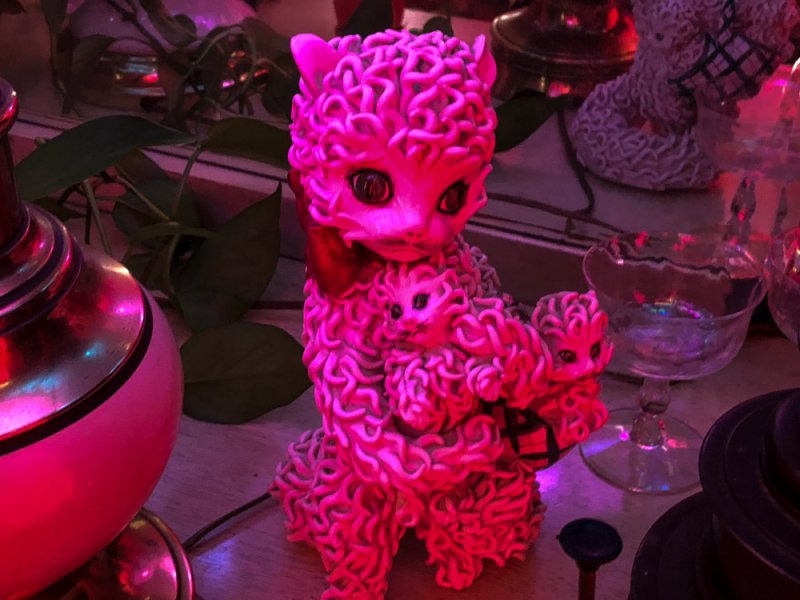 Neon pink cat sculpture with two smaller cats nestled in its curly fur, surrounded by dimly lit objects.