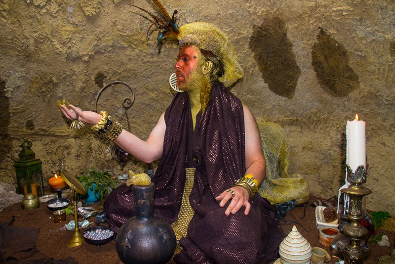 Figure with gold skin, feathered headdress, and brown shawl sits in a cave surrounded by candles, jars, and ritual objects. Earthy tones dominate, with accents of gold and light from the candles.