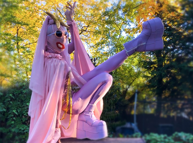 Performance artist in a pink bodysuit, platform boots, and veiled headpiece with horns, posing outdoors.