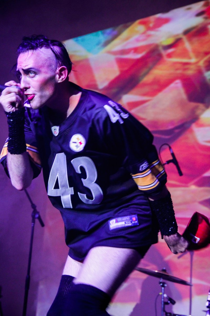 Performance shot: Person in Steelers jersey singing on stage, purple lighting, geometric backdrop.