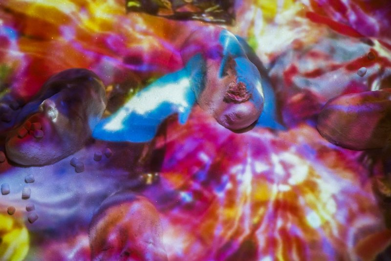 Abstract artwork featuring fleshy forms with blue highlights, scattered sugar cubes, and a single rose. The background is a vibrant mix of reds, yellows, and purples.