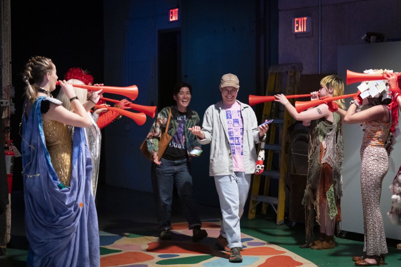 Four performers in costumes blow long red horns at two smiling people walking on a colorful floor.