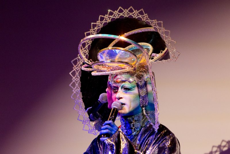 Drag performer Darrell Thorne in elaborate headdress, metallic costume, and dramatic makeup, holding a microphone.