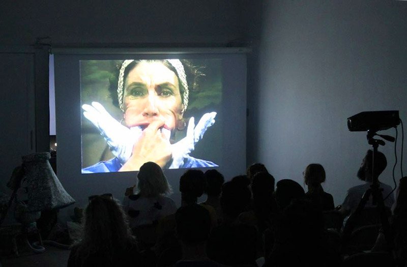 Video projection of a woman with white gloves pulling her mouth, viewed by a dark silhouetted audience in a dim room.