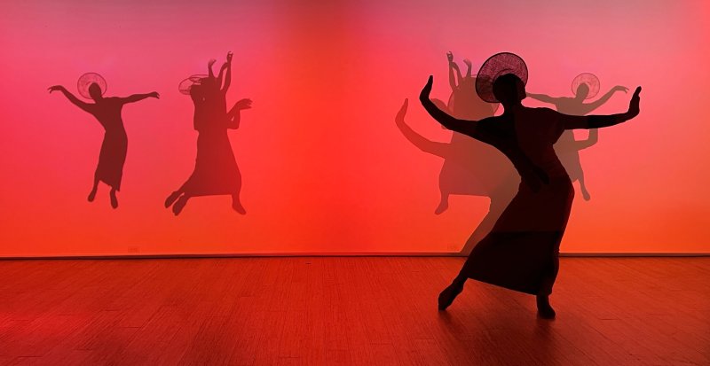 Silhouetted figure in a wide-brimmed hat dancing before a vibrant red and orange gradient wall, with three faint shadow figures behind.