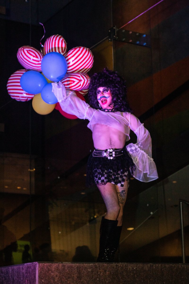 A drag performer with dark curly hair and painted face stands on a platform holding a bouquet of red, white, blue, and yellow balloons. They wear a sheer top, short skirt, boots, and a belt with large decorative buckles.