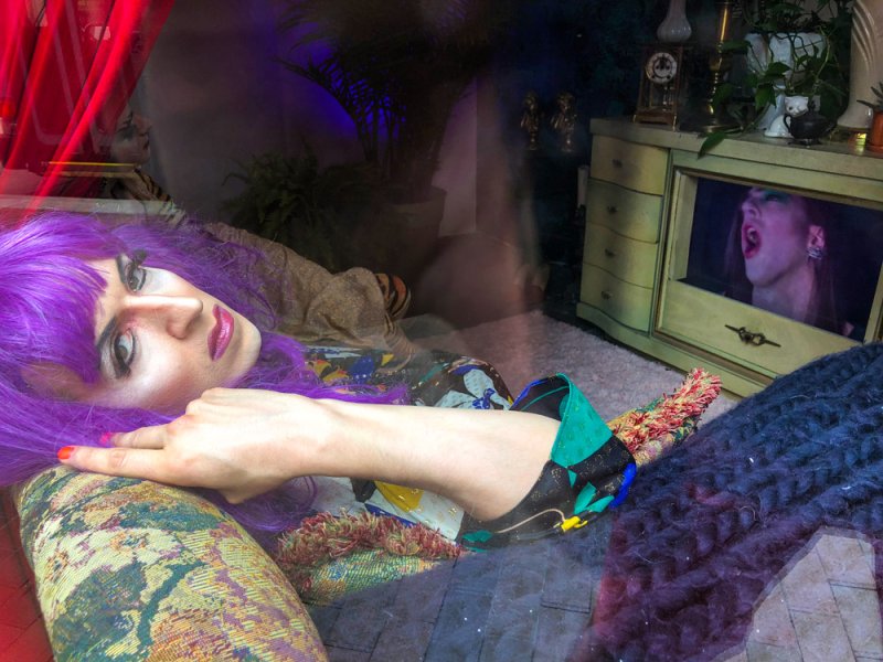 Person with purple hair and dramatic makeup reclines, pointing, with a vintage TV showing a screaming face in the background.
