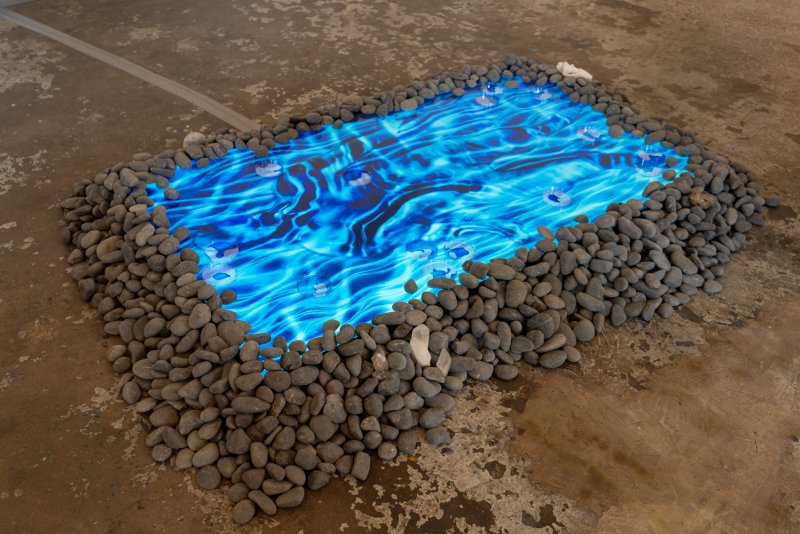 Rectangular pool of rippling blue light on a concrete floor, surrounded by a mound of grey river stones.
