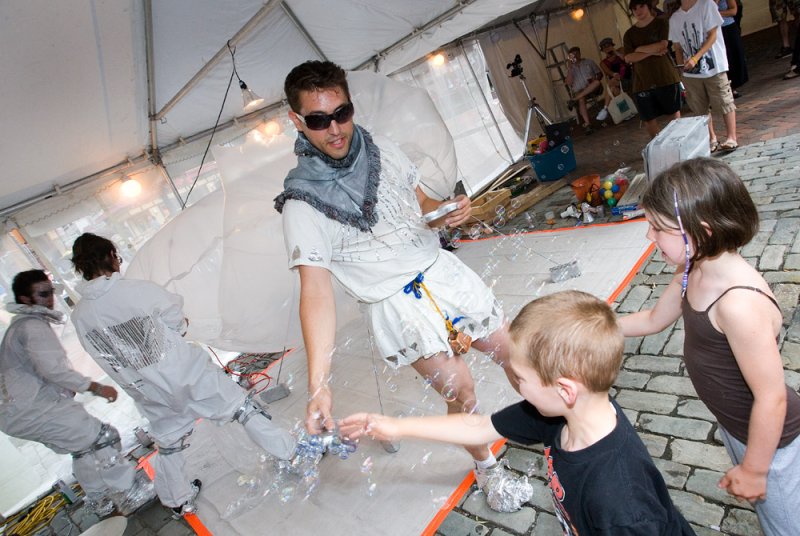 Man in sunglasses and white outfit making bubbles for two children in a tent with other performers.