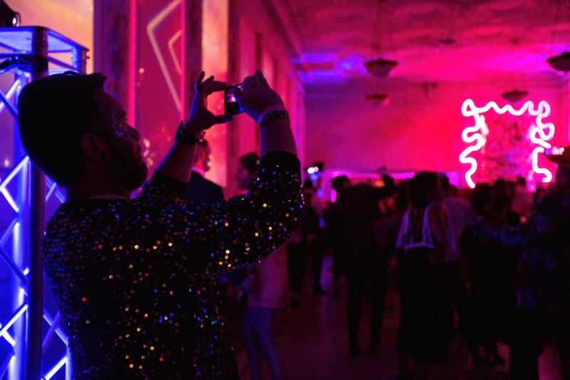 Man in sequined jacket filming a vibrant, neon-lit party with red and blue lighting, blurred figures dancing in background.