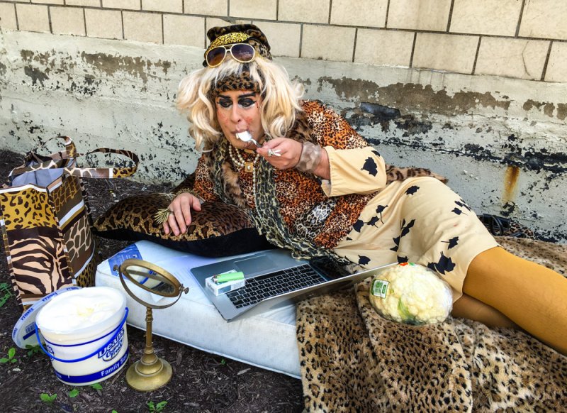 Person in drag reclines on a mattress outdoors, wearing leopard print and sunglasses, eating with a spoon. Laptop, mirror, and bucket of cream nearby.