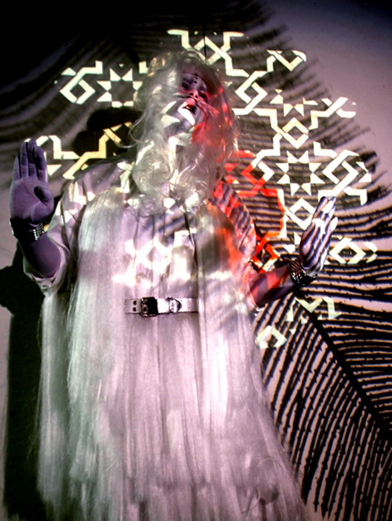 Performance art with a figure in a white wig and garment, hands raised, bathed in projected light patterns and red hues.
