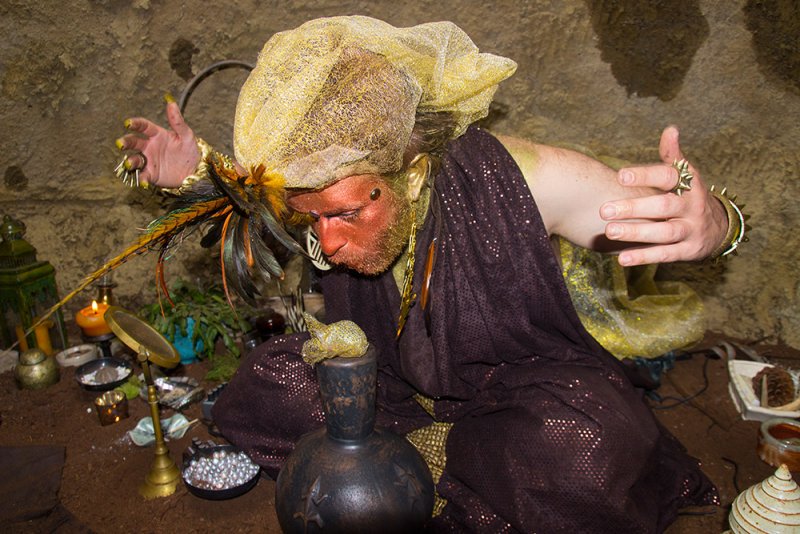 Performance artist in gold makeup and headwrap, brown sequined robe, kneeling before an altar of ritual objects.