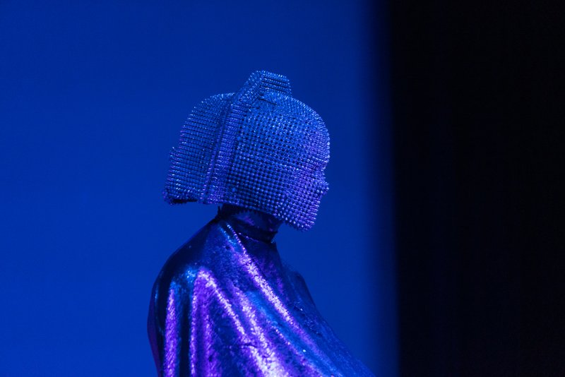 Figure in a shimmering purple garment and a helmet of small, reflective squares, against a blue and dark background.