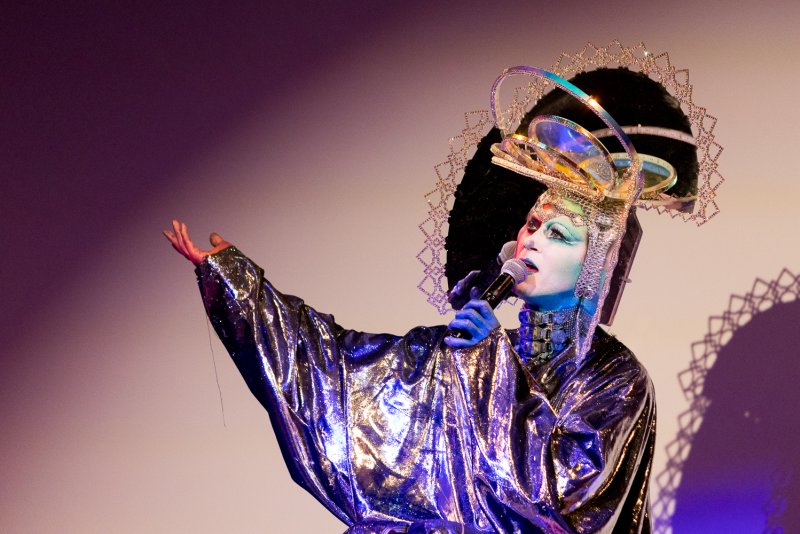 Drag performer Darrell Thorne in a silver robe, elaborate headpiece, and blue-painted skin, singing into a microphone with arm raised.