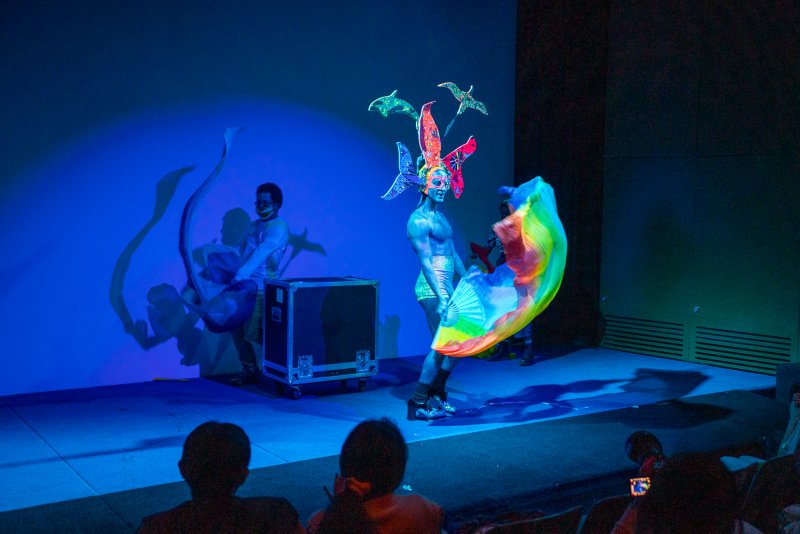 Drag performer Darrell Thorne in a vibrant, feathered headdress and body paint, holding a rainbow fabric on a blue-lit stage.
