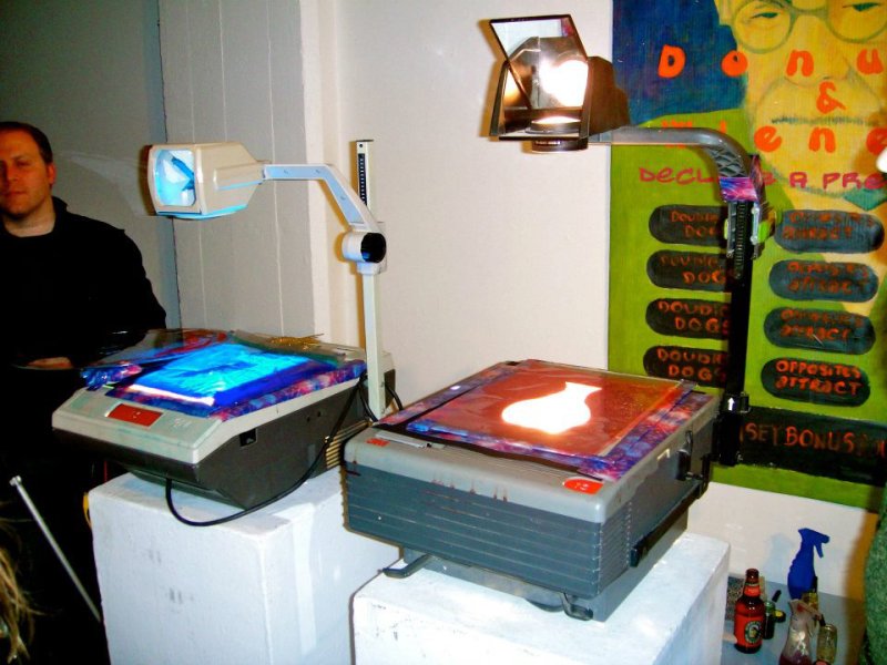 Two overhead projectors display blue and orange liquid light, with a man and a colorful painting in the background.