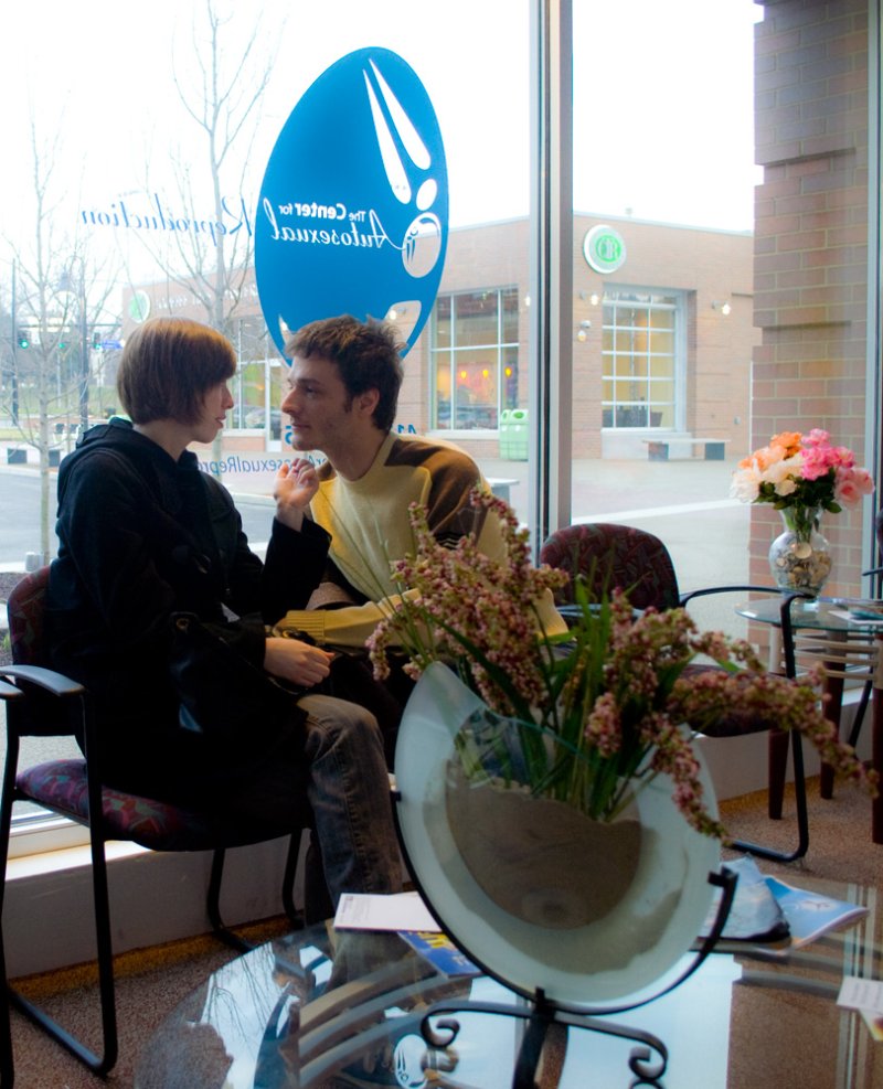 Two people sit facing each other in a waiting room, a vase of flowers in the foreground.