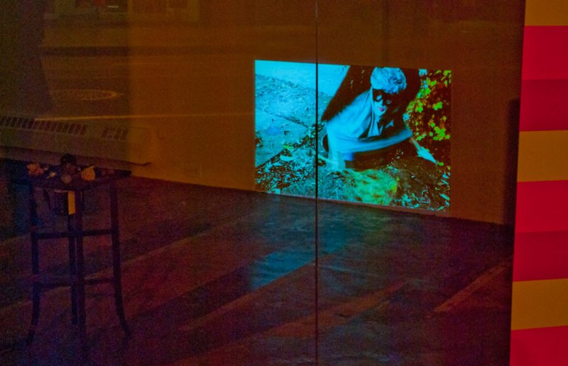 Projected video of a person in a blue-lit outdoor scene, with green foliage, on a gallery wall next to a dark table.