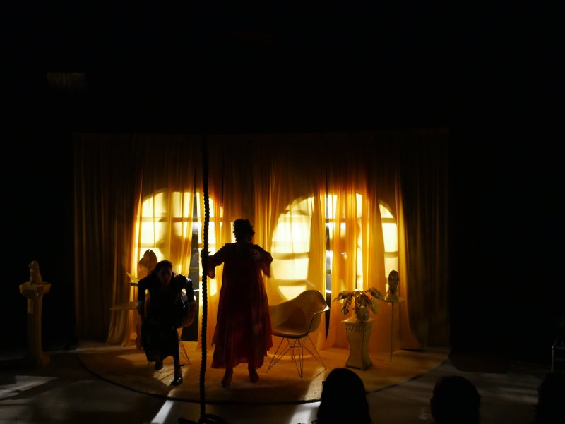 Two silhouetted figures on a circular stage before bright yellow curtains with circular window patterns.