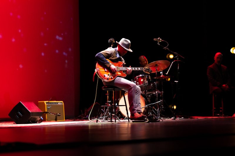 A male guitarist in a hat plays an orange electric guitar on a stage with a red backdrop and drummer.