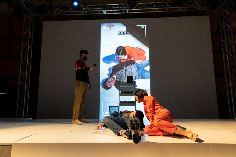 Multi-media installation featuring three figures on a white stage. A large screen projects a video of two people lying down, framed by a camera interface. A person in a mask stands to the left, looking at a phone, while another person in orange sits 